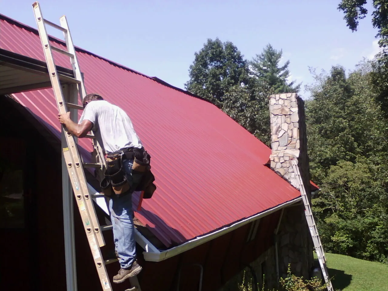 Roofer on a ladder beside a red metal roof and stone chimney — Soffit & Fascia Repair by Roofing Brothers in Brookhaven
